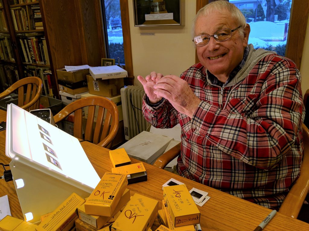 Historian Bill Green sits at a table with boxes of slides and a lightbox in front of him. He holds a slide in his hand as he smiles at the camera.
