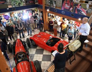 A crowd of people gather in the main hall at the IMRRC awaiting the drawing of the raffle winner. Two red sports cars sit in the center of the hall.